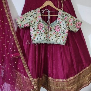 Puede incluir: Una falda de seda magenta con bordados dorados y un dupatta a juego. La falda tiene una cintura fruncida y una silueta acampanada. Se incluye una blusa bordada azul claro con escote en V y mangas cortas.