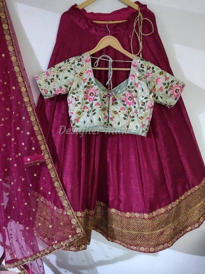 Puede incluir: Una falda de seda magenta con bordados dorados y un dupatta a juego. La falda tiene una cintura fruncida y una silueta acampanada. Se incluye una blusa bordada azul claro con escote en V y mangas cortas.