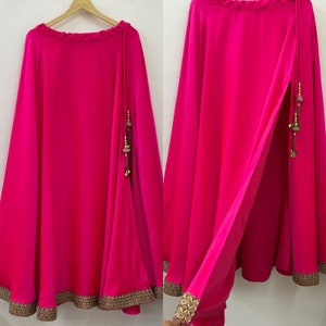 Puede incluir: Una falda maxi de color rosa brillante con un ribete dorado y un largo borla dorado que cuelga del lado. La falda tiene una abertura en el lado.