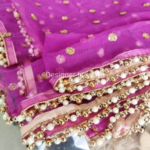 May include: A vibrant magenta net fabric with gold embellishments and a beaded trim. The fabric is adorned with scattered gold sequins and a border of white pearls and gold beads. The design is layered with a light pink fabric.
