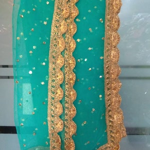 May include: A sheer, teal-colored scarf with gold-colored, scalloped lace trim. The scarf is adorned with small, gold-colored sequins. The lace trim is intricately designed, adding a touch of elegance to the fabric.