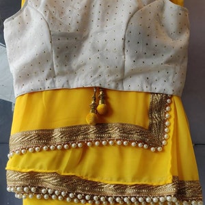 Pu&ograve; includere: Un sari giallo con una blusa bianca ricamata. Il sari ha un bordo dorato e perlato. La blusa ha una scollatura rotonda ed &egrave; senza maniche.