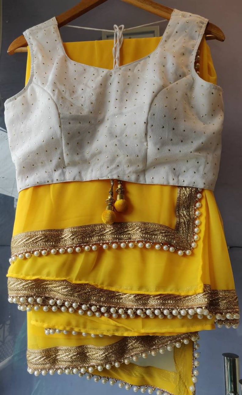 Pu&ograve; includere: Un sari giallo con una blusa bianca ricamata. Il sari ha un bordo dorato e perlato. La blusa ha una scollatura rotonda ed &egrave; senza maniche.