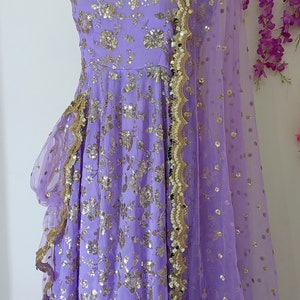 May include: A lavender-colored dress with gold sequins and a matching sheer dupatta. The dress has a ruffled hem and a square neckline. The dupatta has a scalloped border.