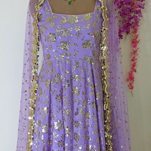 May include: A lavender-colored dress with a floral pattern and gold sequins. The dress has a scoop neckline and a flowing skirt. It is paired with a sheer lavender dupatta with gold trim.