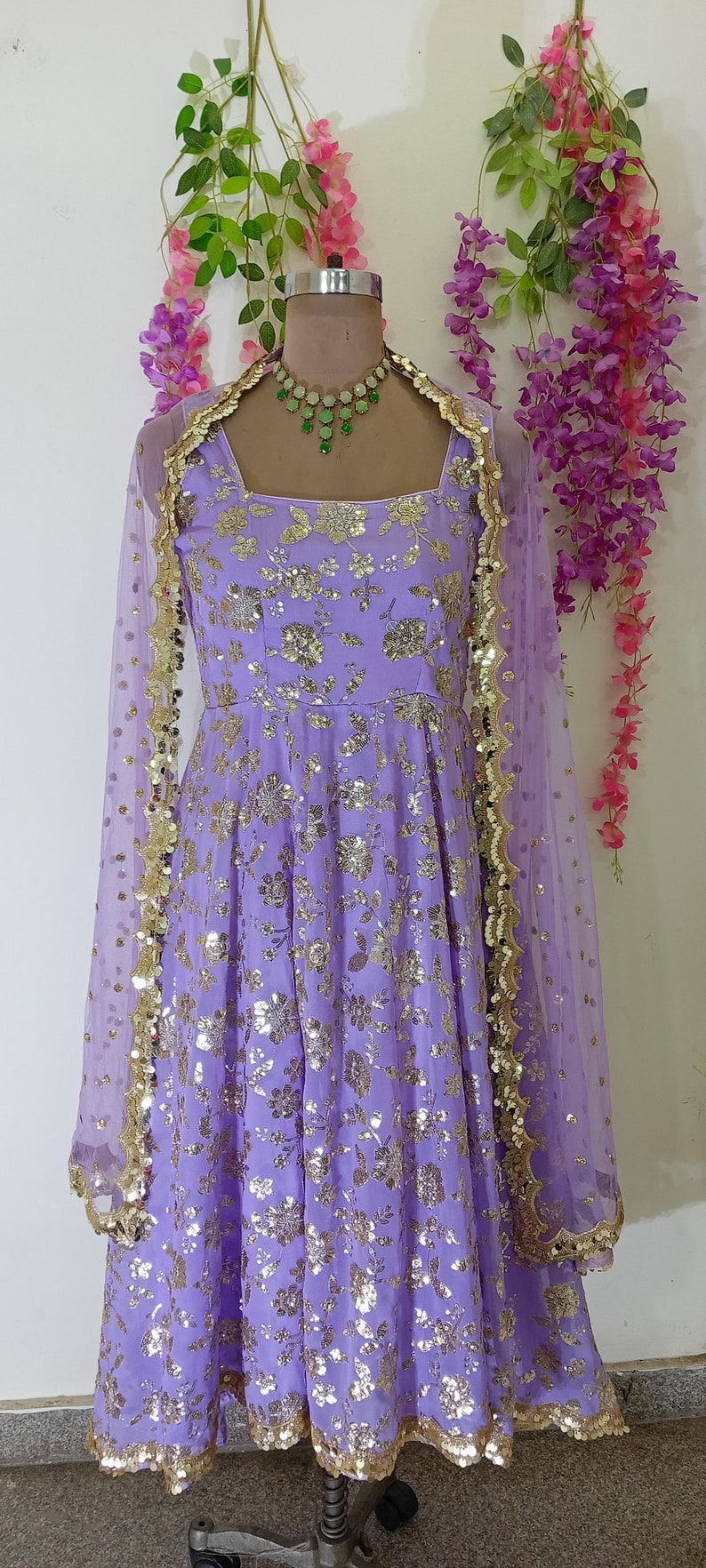 May include: A lavender-colored dress with a floral pattern and gold sequins. The dress has a scoop neckline and a flowing skirt. It is paired with a sheer lavender dupatta with gold trim.