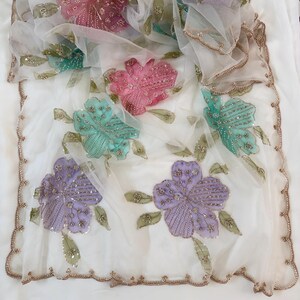 May include: A sheer white fabric with large, embroidered flowers in shades of pink, teal, and purple. The flowers are embellished with sequins and beads. The edges of the fabric are trimmed with a gold-colored, scalloped border.