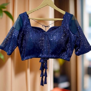 May include: A navy blue sequined top with a square neckline and short sleeves. The top features a sweetheart-shaped bodice and a tie detail at the waist. The sleeves have a sheer mesh panel. The top is hanging on a wooden hanger.