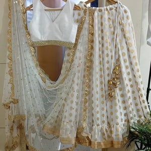 May include: A white, sleeveless top with a V-neck and gold trim, paired with a white skirt featuring gold polka dots and trim. A sheer white scarf with gold trim completes the ensemble. The outfit is hanging on a wooden hanger.