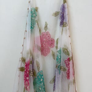 May include: A sheer white fabric with colorful floral embroidery. The flowers are in shades of pink, teal, and purple, embellished with sequins. The edges are trimmed with a delicate gold border.