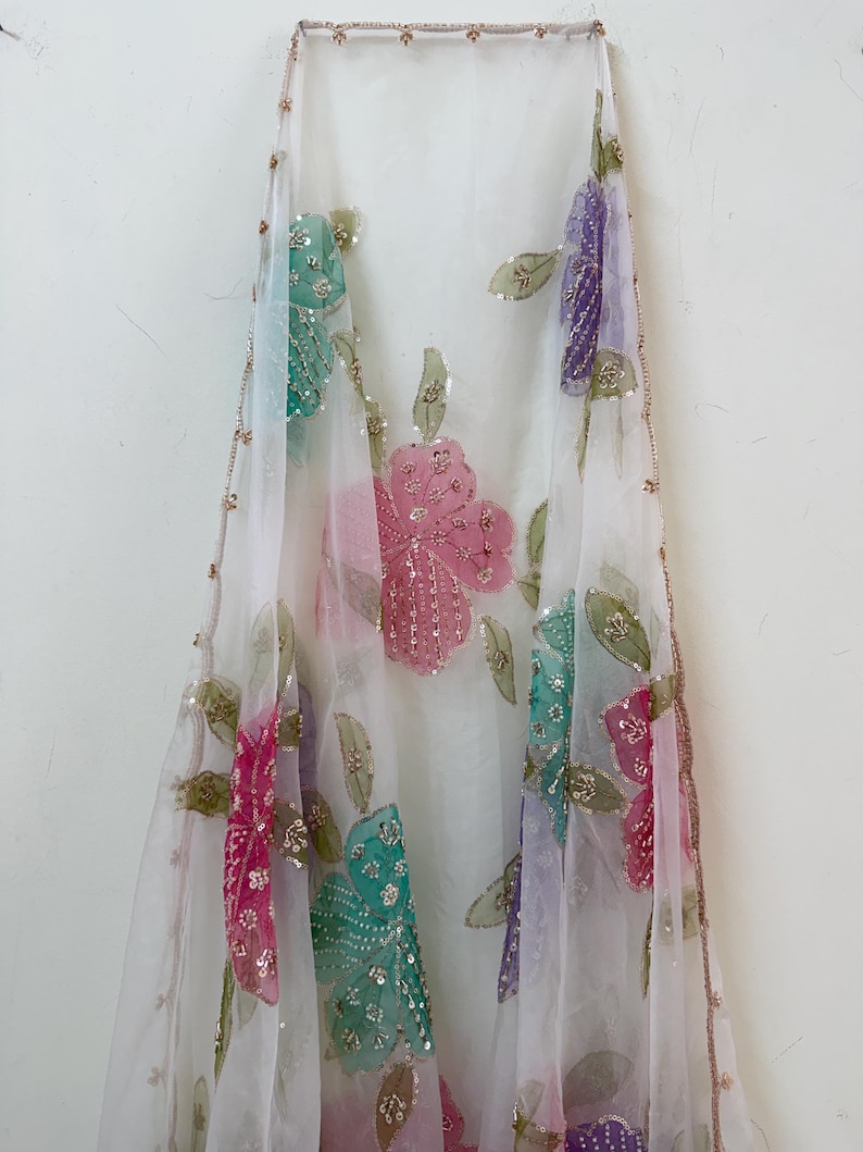 May include: A sheer white fabric with colorful floral embroidery. The flowers are in shades of pink, teal, and purple, embellished with sequins. The edges are trimmed with a delicate gold border.