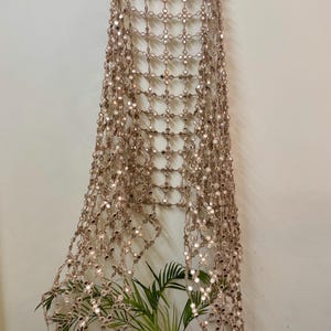 May include: A decorative, open-weave wall hanging with small, reflective mirror accents. The beige-colored piece is suspended from a wooden hanger, creating a grid-like pattern. A potted plant with green leaves is visible at the bottom.