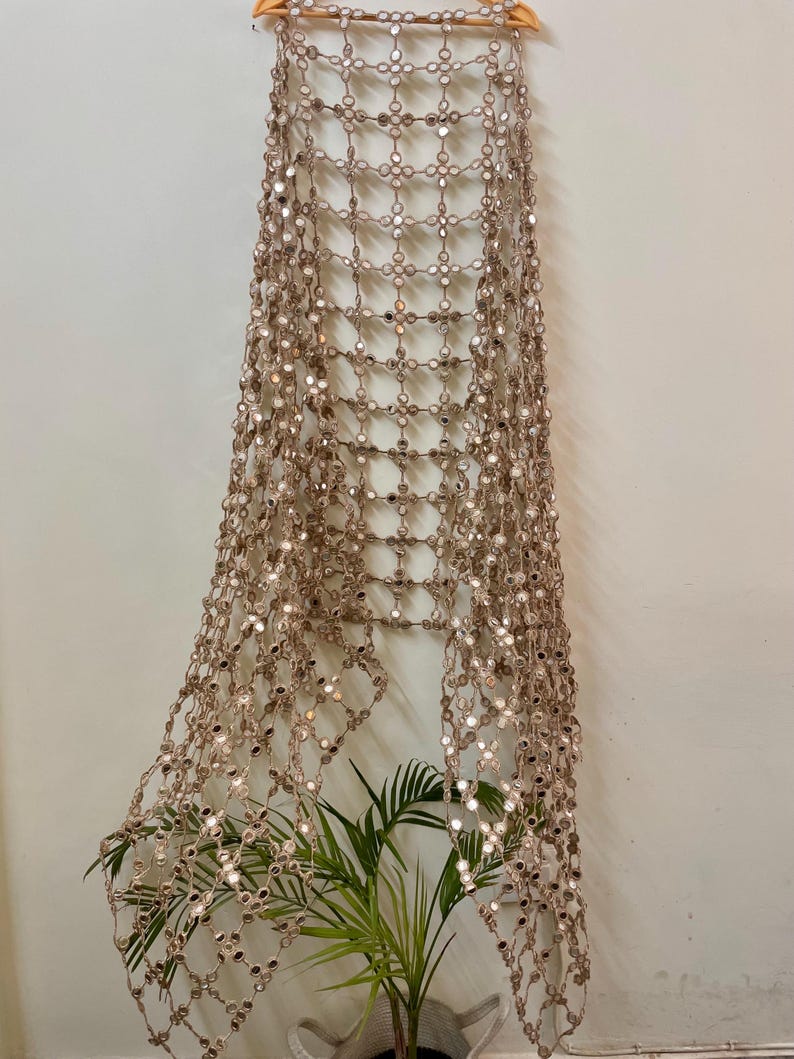 May include: A decorative, open-weave wall hanging with small, reflective mirror accents. The beige-colored piece is suspended from a wooden hanger, creating a grid-like pattern. A potted plant with green leaves is visible at the bottom.