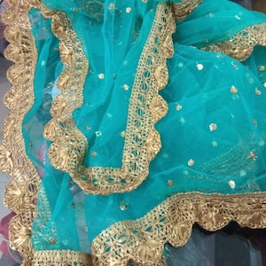 May include: A turquoise sheer fabric with gold-colored decorative trim. The trim has a scalloped edge and intricate floral patterns. Small gold sequins are scattered across the fabric. The fabric is folded, creating a layered effect.