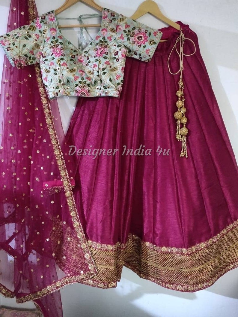 Puede incluir: Una falda de seda magenta con bordados dorados y un dupatta de red a juego. La falda tiene un cierre de cord&oacute;n y una borla dorada. La blusa es verde claro con bordados florales.