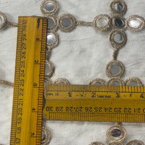 May include: Close-up of gold-colored trim with small, round mirrors. A yellow ruler shows measurements in inches. The trim is on a white fabric background. The ruler shows measurements up to 4 inches.