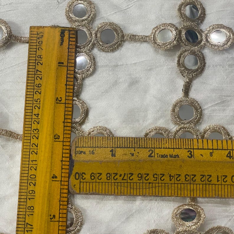 May include: Close-up of gold-colored trim with small, round mirrors. A yellow ruler shows measurements in inches. The trim is on a white fabric background. The ruler shows measurements up to 4 inches.