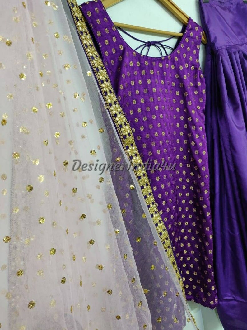 Op de afbeelding: Een paarse en gouden geborduurde jurk met een witte net dupatta. De jurk heeft een ronde halslijn en is mouwloos. De dupatta heeft gouden pailletten.