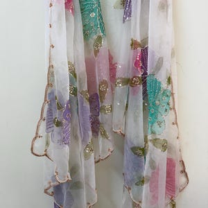 May include: A sheer white scarf with colorful floral embroidery. The scarf features pink, green, purple, and teal flowers with gold sequin accents. The edges are scalloped and trimmed with gold thread, creating a delicate and elegant look.