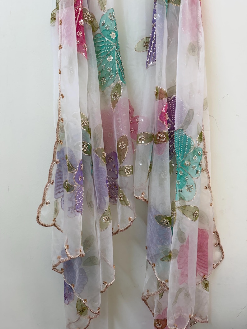 May include: A sheer white scarf with colorful floral embroidery. The scarf features pink, green, purple, and teal flowers with gold sequin accents. The edges are scalloped and trimmed with gold thread, creating a delicate and elegant look.