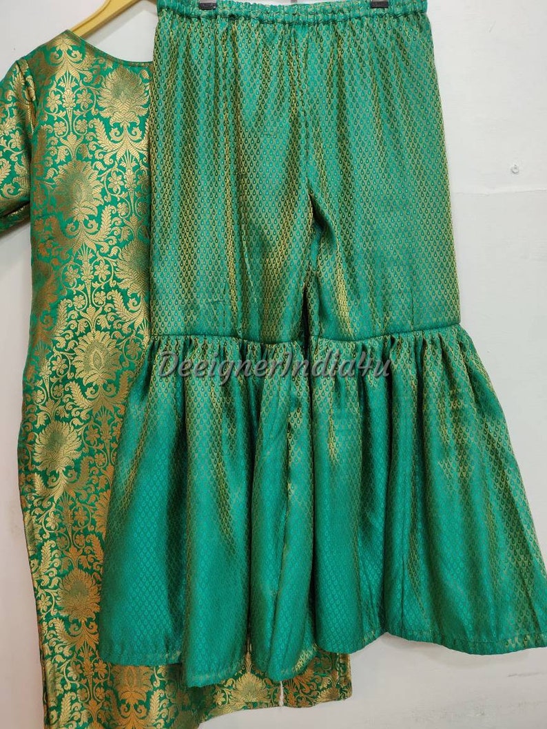 May include: A green and gold brocade outfit, including a top and flared pants. The fabric has a detailed floral pattern. The pants have a ruffled hem. The outfit is displayed on a hanger.