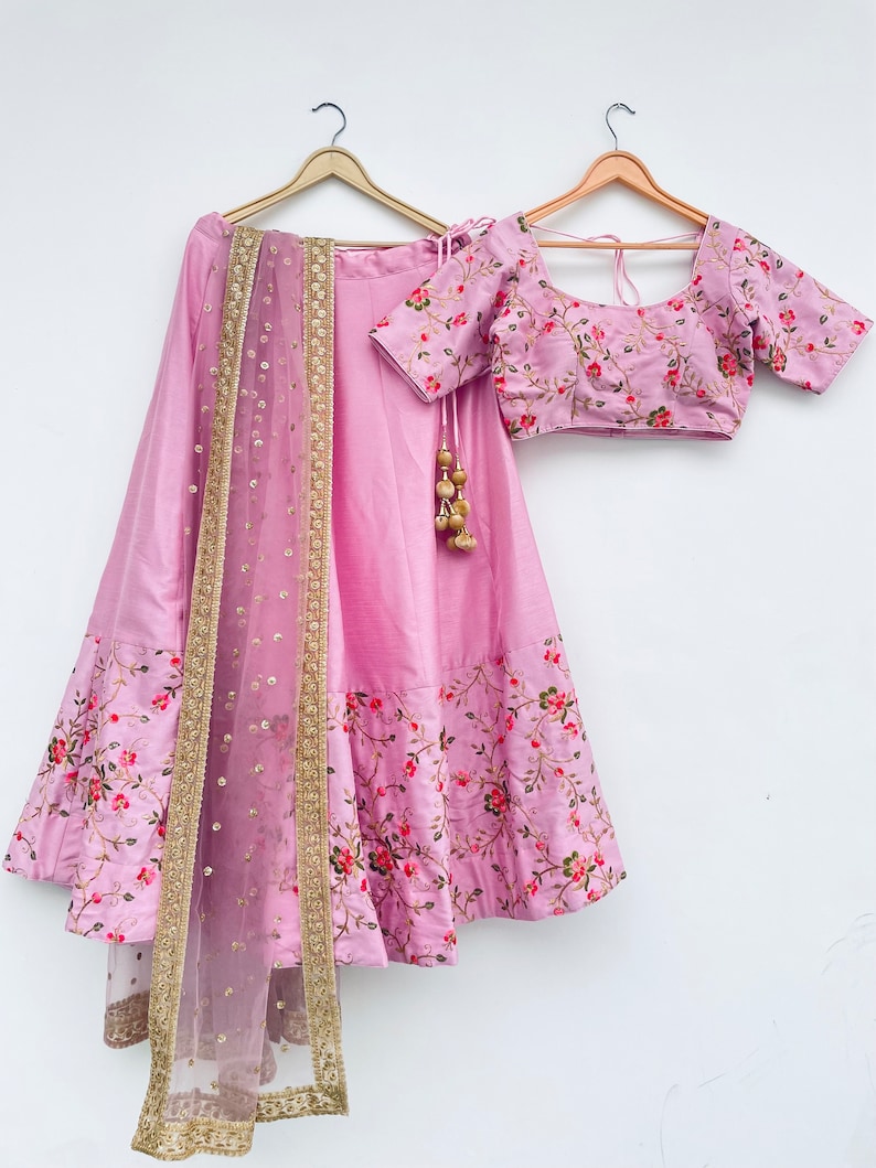 May include: A pink and floral embroidered lehenga with a matching pink embroidered blouse and a sheer pink dupatta with gold trim.
