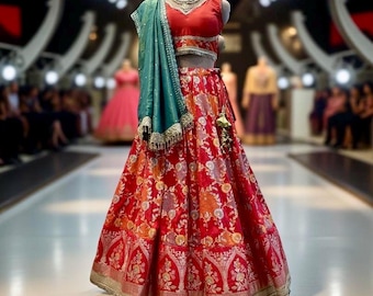 May include: A red and gold embroidered lehenga with a teal dupatta. The lehenga has a floral pattern and a gold border. The dupatta is draped over the shoulders and has a gold tassel.