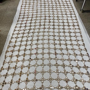 May include: A large, rectangular textile featuring a grid-like pattern of small, round mirrors. The mirrors are connected by a network of tan-colored threads, creating a decorative, openwork design. The textile is laid out on a white surface.