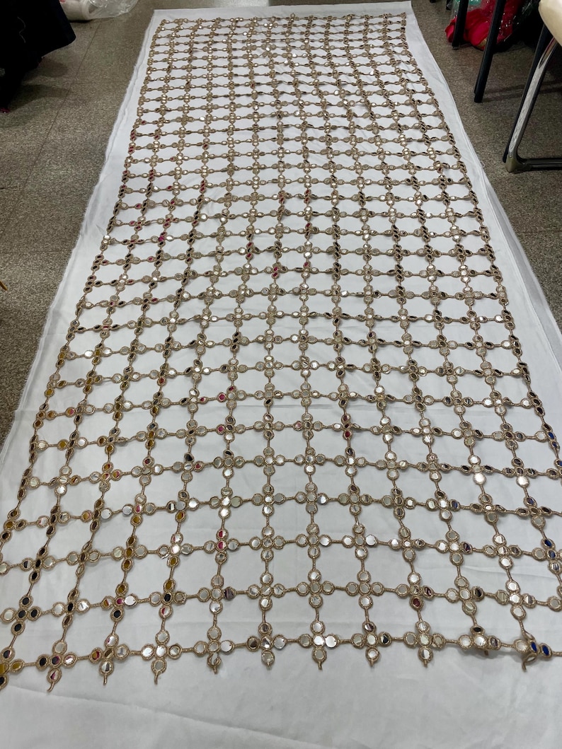 May include: A large, rectangular textile featuring a grid-like pattern of small, round mirrors. The mirrors are connected by a network of tan-colored threads, creating a decorative, openwork design. The textile is laid out on a white surface.