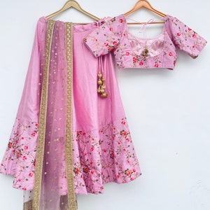 May include: A pink and gold embroidered lehenga with a matching pink and gold embroidered blouse. The lehenga has a floral print and a net dupatta with gold sequins.