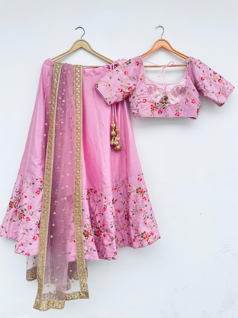 May include: A pink and gold embroidered lehenga with a matching pink and gold embroidered blouse. The lehenga has a floral print and a net dupatta with gold sequins.