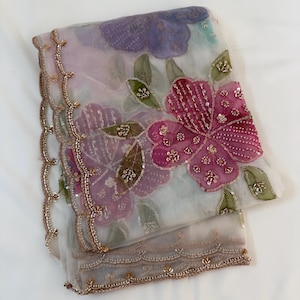 May include: A sheer, light-colored fabric with floral embroidery. The design features large, pink and purple flowers with sequin details and green leaves. The edges are trimmed with a scalloped border and gold beading.