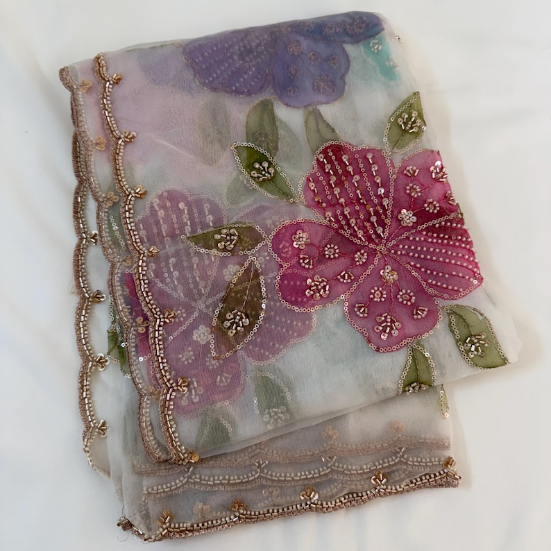 May include: A sheer, light-colored fabric with floral embroidery. The design features large, pink and purple flowers with sequin details and green leaves. The edges are trimmed with a scalloped border and gold beading.