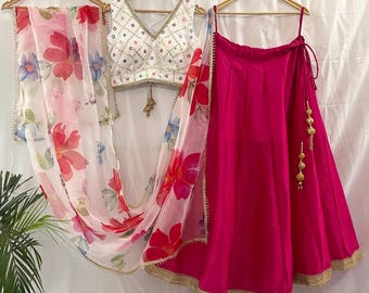 May include: A white crop top with a geometric design and a pink skirt with gold trim. The skirt has a tie at the waist and a tassel on the side. The outfit is completed with a sheer white dupatta with floral print.