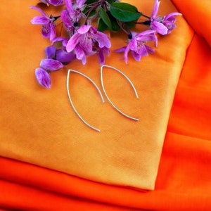 May include: A pair of silver earrings with a simple, curved design. The earrings are displayed on a bright orange and peach fabric background with purple flowers.