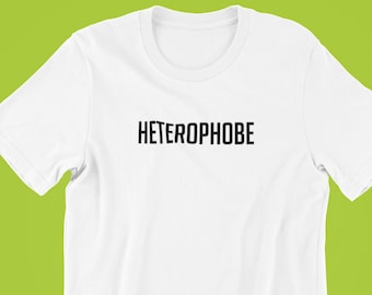 以下が含まれることがあります： 中央に黒字で「HETEROPHOBE」とプリントされた白い半袖Tシャツ。柔らかい素材で作られたシャツは、緑色の背景に表示されています。