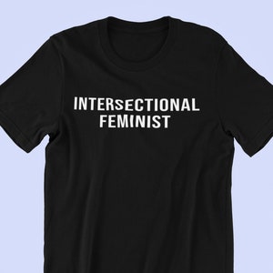 Puede incluir: Camiseta negra con el texto blanco "INTERSECTIONAL FEMINIST" en el pecho. La camiseta de manga corta está hecha de un material suave y se muestra sobre un fondo azul claro.