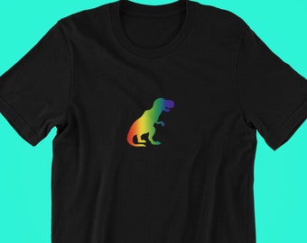 May include: Black t-shirt featuring a rainbow-colored Tyrannosaurus Rex graphic. The dinosaur design is a gradient of colors, including red, orange, yellow, green, blue, and purple. The shirt is made of a soft material.