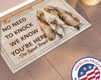 May include: A welcome mat with a brown basset hound illustration and the text "No need to knock we know you're here - The Basset Hound". The mat has a beige background with a brown border.