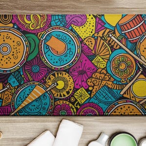 May include: A rectangular bath mat with a vibrant, colorful design. The pattern features stylized drums, flowers, and geometric shapes in shades of orange, teal, pink, yellow, and black. The mat is placed on a light wood surface.