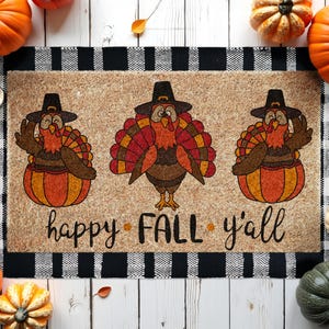 May include: A rectangular doormat with a tan background features three cartoon turkeys wearing pilgrim hats and sitting in pumpkins. The text "happy FALL y'all" is written in black script. The mat has a black and white checkered border.