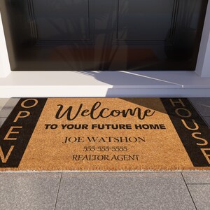 Op de afbeelding: Een bruine kokosmat met zwarte letters die "OPEN HOUSE" aan de linker- en rechterkant aangeven. Het midden van de mat geeft "Welcome to your future home" aan met de naam "Joe Watson" en een telefoonnummer eronder. De tekst "Realtor Agent" staat onderaan.