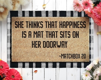 May include: A rectangular doormat with the text "SHE THINKS THAT HAPPINESS IS A MAT THAT SITS ON HER DOORWAY -MATCHBOX 20" in black. The mat is brown with a black and white checkered border.