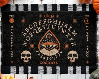 Puede incluir: Felpudo negro con diseño de tablero Ouija. La alfombra presenta el alfabeto, números y las palabras "Sí", "No" y "Adiós". Incluye un símbolo de ojo central y gráficos de calaveras, con un borde a rayas blanco y negro.