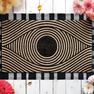 May include: Rectangular doormat with a geometric eye design. The mat features concentric circles and curved lines in shades of brown and black. A black and white striped border frames the mat.