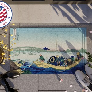 May include: A blue and white doormat with a Japanese woodblock print of a boat with people on it. The print is of a scene with a bridge and a mountain in the background. The doormat is made in the USA.