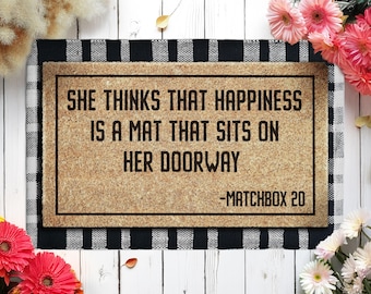 May include: A rectangular doormat with the text "SHE THINKS THAT HAPPINESS IS A MAT THAT SITS ON HER DOORWAY -MATCHBOX 20" in black lettering. The mat has a brown surface and a black and white checkered border.
