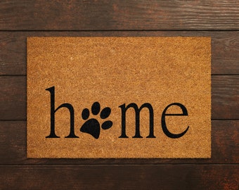 May include: A brown coir doormat with the word "home" in black lettering and a black paw print between the "h" and the "me".