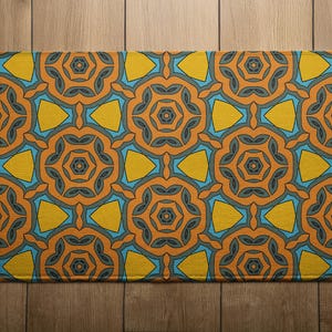May include: Rectangular rug with a repeating geometric pattern. The design features orange, yellow, and blue shapes on a textured background. The rug is placed on a wooden floor.