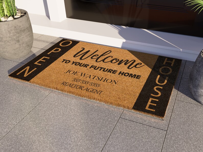 Op de afbeelding: Een bruine kokosvezel welkommat met zwarte letters die "Open House" en "Welcome to your future home" zeggen. De mat bevat ook de naam, het telefoonnummer en de titel van een makelaar: "Joe Watshon, 555-555-5555, Realtor Agent."